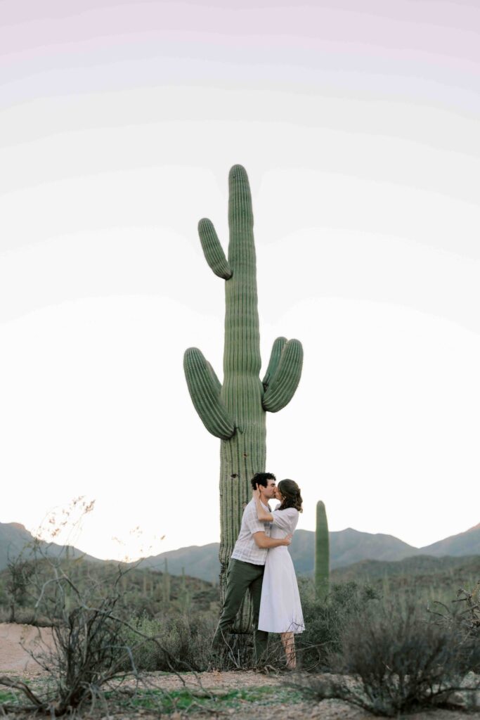 An engaged couple kisses in front of a large cactus during an Arizona sunset for an engagement photo.