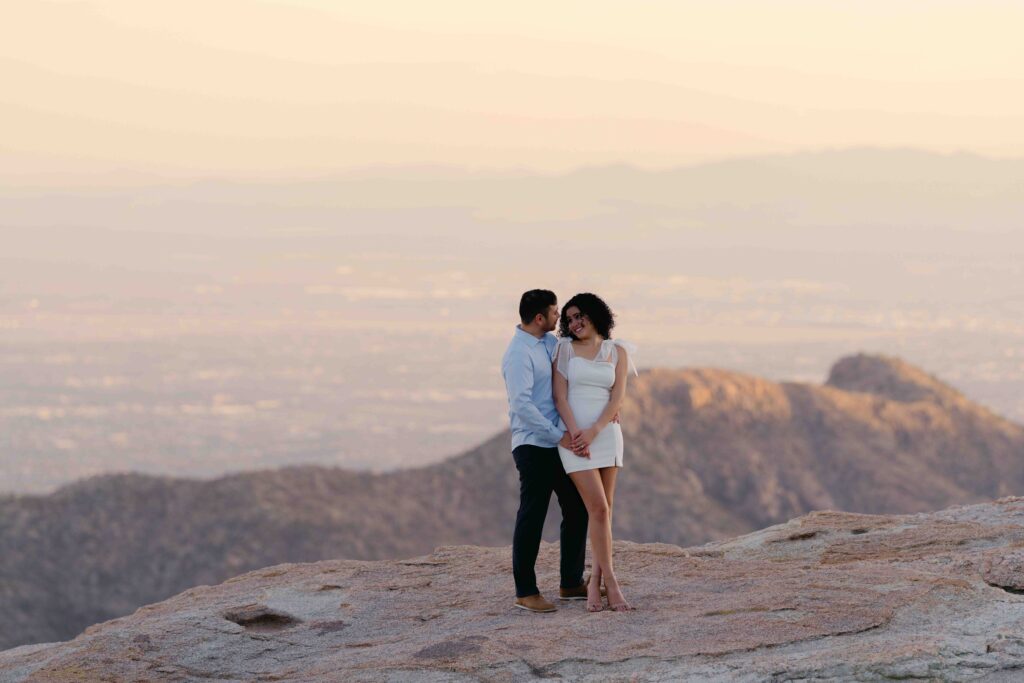 A couple stands on a mountain during their engagement session just west of Phoenix Arizona.  The bride wears a short white dress for their engagement session and the groom wears a blue shirt with black slacks. 