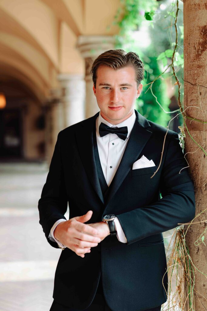 A groom in black tie attire stands against a pillar covered in vines looking at the camera at his Arizona wedding. 
