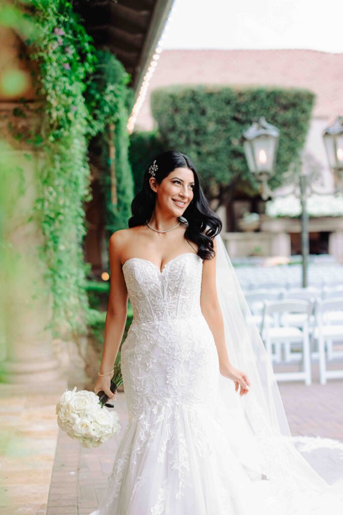 A bride smiles and looks out over her ceremony space at Vila Siena in Arizona.  She's wearing a lace dress and holding a bouquet of white roses. 
