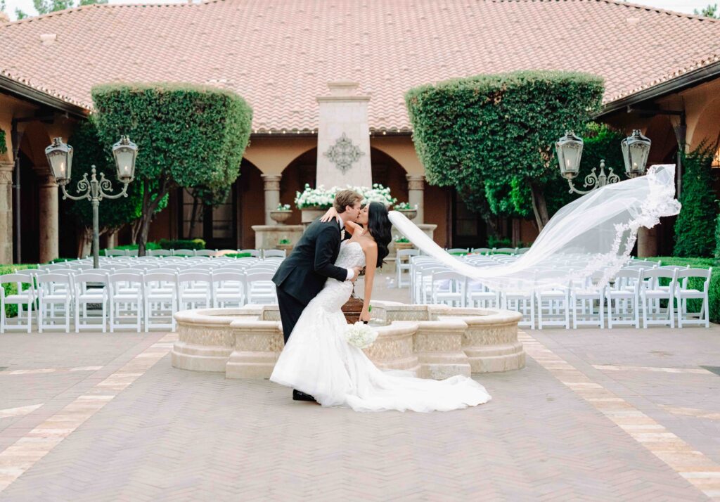A bride wearing a lace trimmed cathedral veil kisses her husband during their wedding day in a courtyard at Villa Siena.  