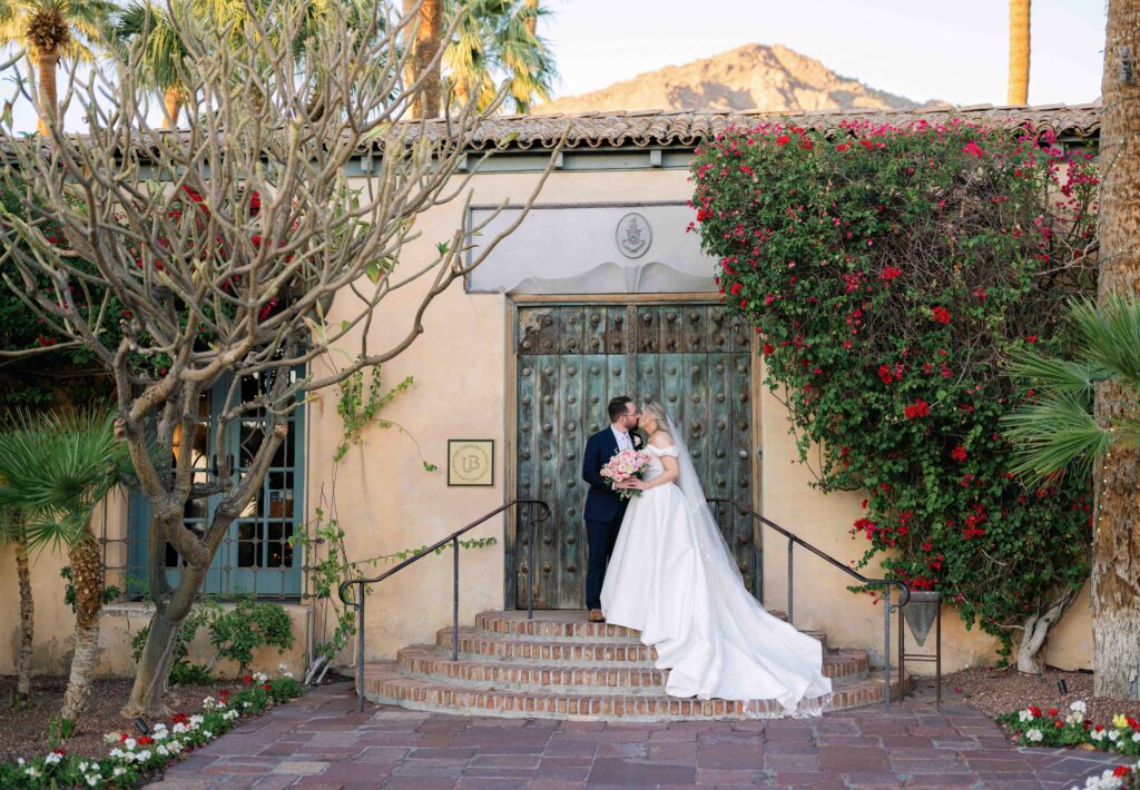 A bride and groom stand outside a beautiful antiqued door at their outdoor wedding venue, Royal Palms in Scottsdale Arizona.  